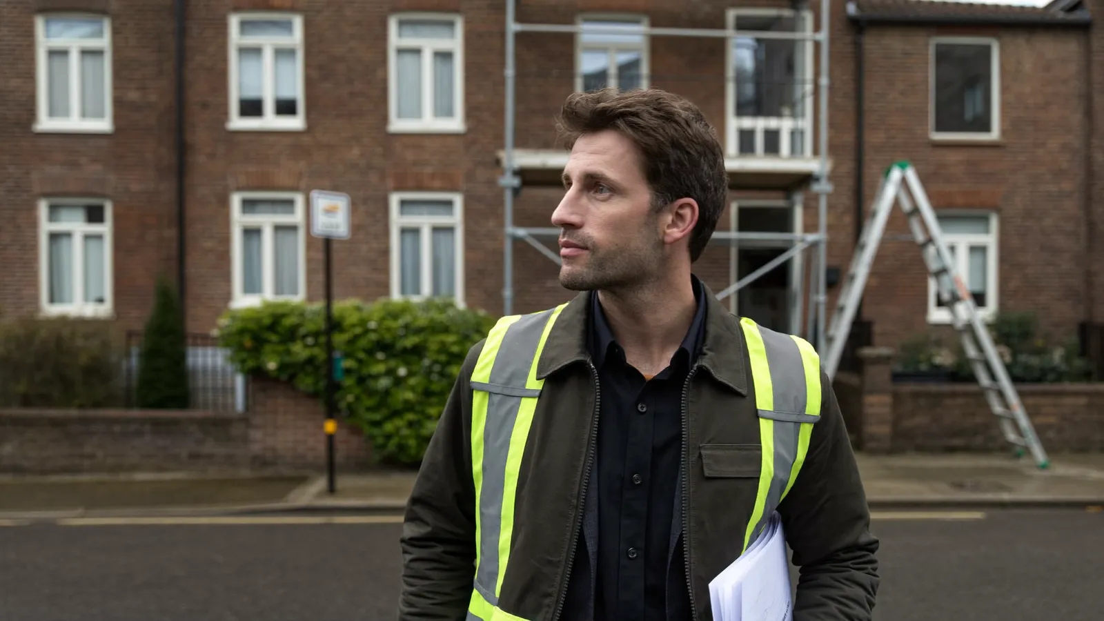 Aannemer met een dossiermap voor een bakstenen rijwoning in renovatie, met stelling en ladder tegen de gevel